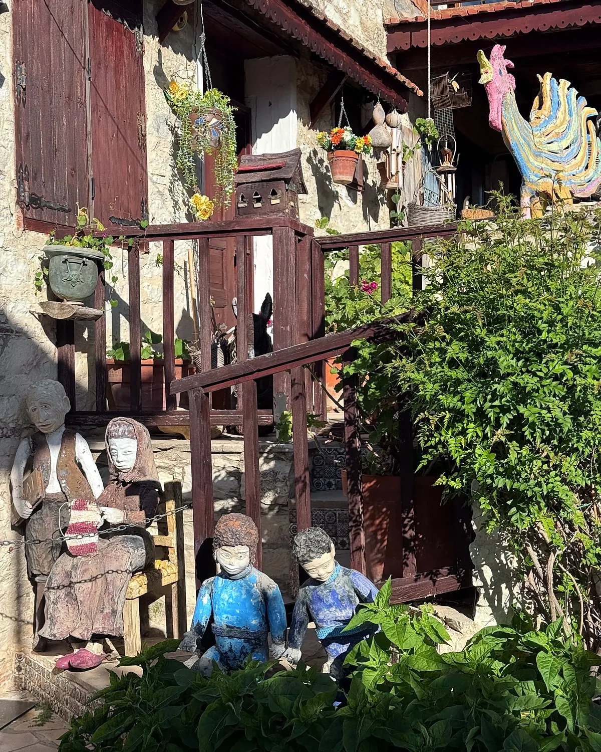 Lania village, Cyprus: we walked into every open door Painted figures sitting by a wooden stairway with plants and a colourful rooster in a Lania courtyard