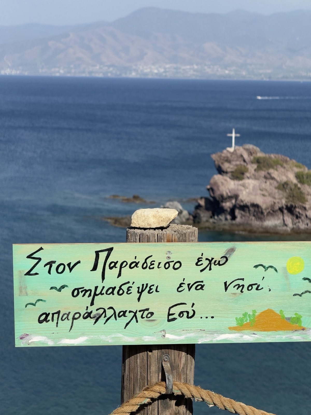 Hand-painted sign in Greek by the shore, with the rock with the cross beyond
