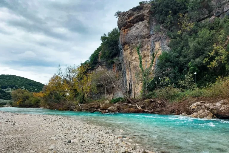The turquoise Acheron River flowing past a limestone cliff and pebble bank in Epirus, Greece, under an autumn sky