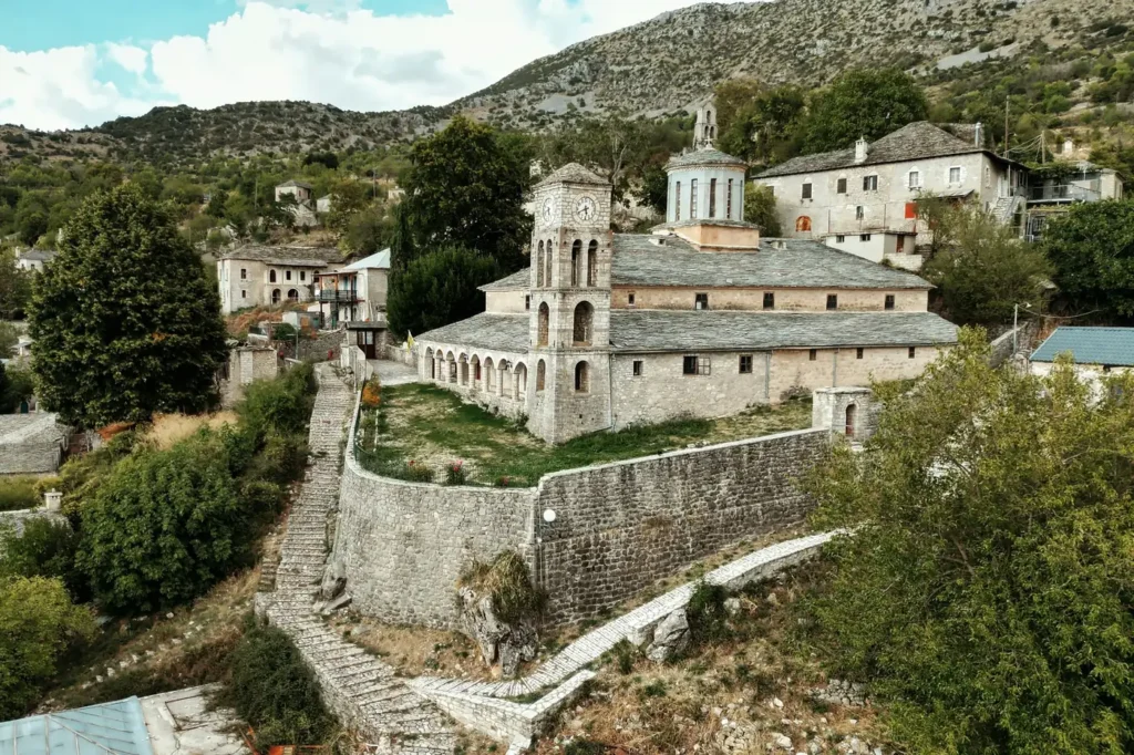 Destinations Stone church with a clock tower and slate roofs above a walled courtyard, with stone steps climbing the hill in Kalarrytes
