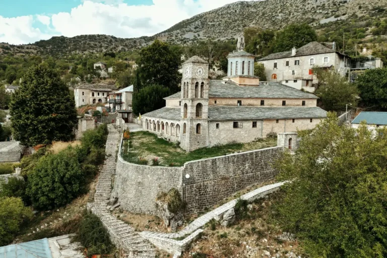 Stone church with a clock tower and slate roofs above a walled courtyard, with stone steps climbing the hill in Kalarrytes