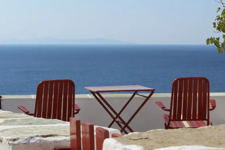 Two red wooden chairs and a small table on a whitewashed terrace overlooking the Aegean Sea in Sifnos
